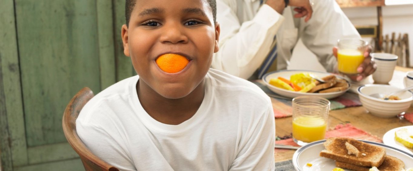 Boy at the breakfast table with an orange in his mouth