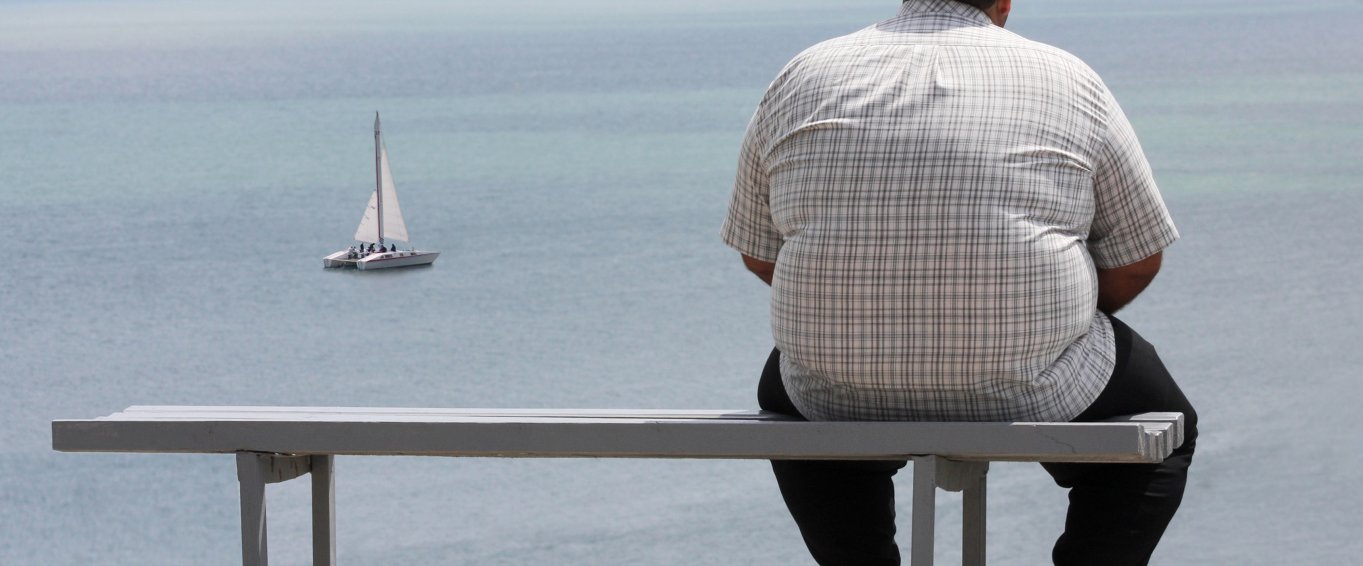 Obese man sitting on a bench, looking at a boat