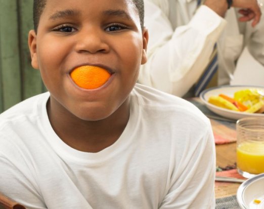 Boy at the breakfast table with an orange in his mouth