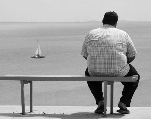 Obese man sitting on a bench, looking at a boat